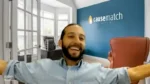 Smiling man with a beard and light blue shirt raises his arms in an office, with a blue wall displaying the 'causematch' logo in the background.