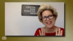 Smiling woman with short blonde hair and glasses on a video call, in front of a sign that reads Center for Positive Media, wearing a red top and gesturing with hands up.