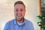 Smiling man with blond hair and beard in a blue checkered shirt, seated in an office with a whiteboard behind him and a plant to the right.