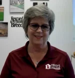 Smiling woman with short gray hair and glasses wearing a maroon polo with the Heidi's Village logo, in an office setting with wall art behind her.