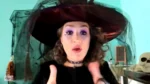 Woman wearing a large witch hat and purple makeup speaks to the camera in a teal-lit room, Halloween decor in the background.