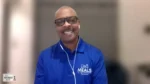 Smiling man in a blue polo with a Meals on Wheels logo, wearing white earbuds during a video call (The Nonprofit Show watermark bottom-left).