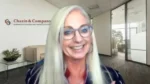 Smiling woman with long gray hair and glasses in an office lobby with the Chazin & Company logo in the background.