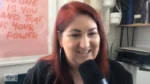 Smiling woman with red hair speaks into a microphone in a studio, pink motivational poster in the background.