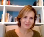 Smiling woman with light brown hair in a brown sweater, seated on a beige chair with bookshelves in the background.