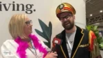 Interviewer with pink feather boa interviewing a man in a bright ceremonial hat and fringed jacket at an expo.
