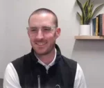 Smiling man with short hair and glasses, wearing a white shirt and black vest, seated indoors with a plant and shelf behind him.