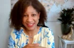 Smiling woman with curly hair in a blue floral blouse, seated indoors with a map on the wall behind her.