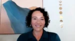 Smiling woman with short curly black hair on a video call, wearing a dark teal shirt, with abstract art in the background.