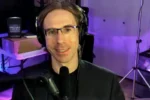 Man with glasses and headphones speaks into a microphone in a recording studio with purple lighting in the background.