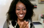 Smiling woman with curly hair claps her hands at a desk in a bright home office.
