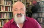 Smiling bald man with a white beard in a home library, with shelves of books and hats hanging in the background.