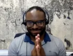 Man wearing glasses and a headset sits at a desk, hands pressed together in a Namaste greeting on a video call.