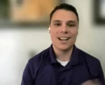 Smiling man in a purple polo shirt during a video call, wearing wireless earbuds with a blurred background.