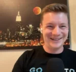 Smiling man with short hair and a wireless earbud speaks into a microphone, with a city night skyline wall art featuring a red moon in the background.