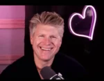 Smiling man hosting a podcast, seated at a microphone with a pink neon heart in the background.