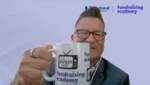 Smiling man in a suit and glasses holds a white mug that reads 'NONPROFIT SHOW' with a TV icon; backdrop shows 'fundraising academy' branding.
