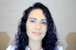 Close-up portrait of a woman with dark, curly hair and pink lips, looking at the camera against a light geometric wall backdrop.