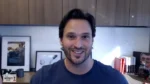 Smiling man in a blue T-shirt on a video call, with framed photos on a desk and wooden cabinets behind him.
