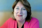 Smiling woman with short light brown hair wearing a pink cardigan on a video call.