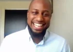 Smiling man with a beard in a light blue shirt during a video call.