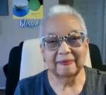 Close-up of an older woman with short white hair wearing patterned glasses and a dark shirt, smiling at the camera in a home office setting.