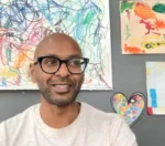 Man with glasses smiling, in a light gray shirt, in front of colorful abstract wall art and a heart-shaped mosaic sign behind him.