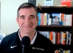 Smiling man with short dark hair speaking into a microphone, with bookshelves in a study or office behind him.