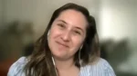 Woman with brown hair smiling at the camera during a video call, wearing white earphones and a light blue striped blouse.