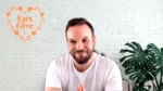 Smiling man speaks to camera in a bright room with a white brick wall and an orange Lux Give heart logo behind him.