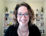 Smiling woman with curly hair and glasses on a video call, in a bright home office with bookshelves behind her.