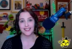 Smiling woman in a home studio with a large blue microphone, green LED lights, and framed art behind her.