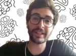 Man with dark hair and glasses speaks during a video call, wearing a dark shirt with white earphones, background has abstract black doodles on white wall.