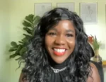 Close-up of a smiling Black woman with wavy black hair, wearing a black top and silver necklace, in a bright room with plants in the background.