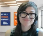 Smiling woman with black glasses and dark hair, wearing a plaid top, in an office with blue and orange posters that say 'We build stronger nonprofits'.