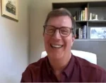 Smiling man wearing glasses and a maroon shirt during a video call in a home office with a bookshelf in the background.