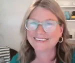 Smiling woman with shoulder-length light brown hair and large round glasses, wearing a teal top, indoors with a shelf in the background.