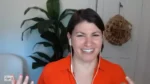Smiling woman on a video call wearing an orange top and white earbuds, with a plant and paper decoration in the background.