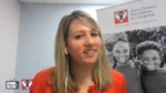Woman in a red top speaks on a video call, wearing earbuds, with a Dave Thomas Foundation for Adoption banner in the background.