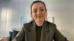 Woman smiling at the camera during a video call, wearing a dark green blazer in a light-paneled room with framed art behind her.