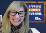 Smiling woman with blonde hair and blue glasses in front of a sign that says 'Building Stronger Nonprofits'.