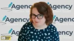 Woman with glasses seated in front of a branded backdrop reading 'Agency Content Writer' repeatedly; appears in a video call setup.
