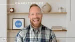 Man in a plaid shirt smiling during a webinar, wearing wireless earbuds with a home office backdrop and CharityBuzz branding above him.
