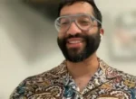 Man with a beard and large clear glasses smiling at the camera during a video call, wearing a colorful patterned shirt and wireless earbuds.