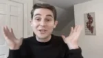 Young man in a black sweater gestures with both hands raised while talking to the camera in a home setting.