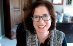 Smiling woman with short brown hair and glasses on a video call, indoor office-like room in background.