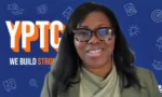 Smiling woman wearing glasses on a video call, with a blue YPTC background reading 'YPTC' and 'We build strong'.