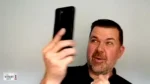 Man filming himself with a smartphone held up in front of a light wall, logo of 'The Nonprofit Show' in the bottom-left corner