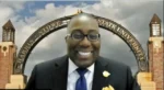Smiling man in a dark suit and blue tie posing in front of a Harris-Stowe State University arch backdrop.
