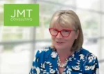 Smiling woman with red glasses and pearl necklace in a blue floral blouse, in an office setting; JMT Consulting logo in a green box top-left.
